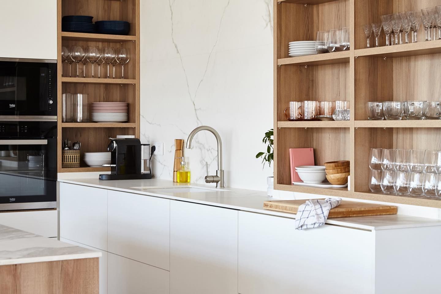Cocina moderna blanca con estantes de madera, grifo de acero inoxidable y horno empotrado.
