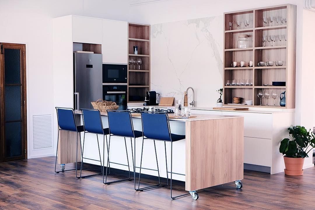 Cocina moderna con isla, gabinetes blancos, detalles de madera, taburetes de bar azules y estantes abiertos.