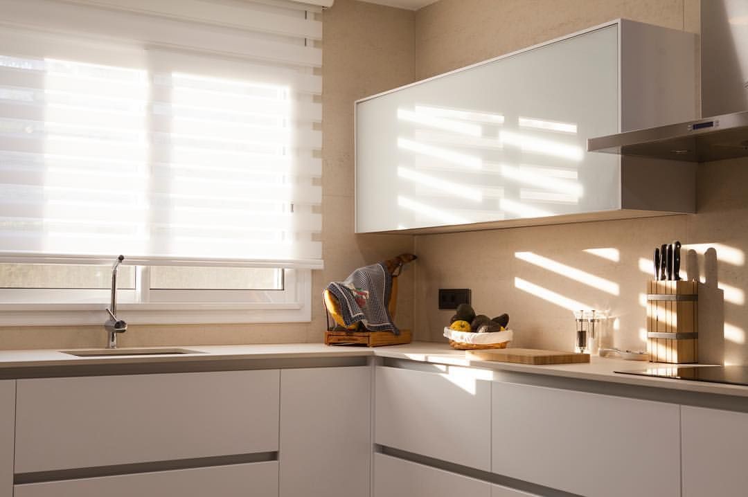 Cocina moderna con gabinetes blancos, ventana con persianas y luz solar.