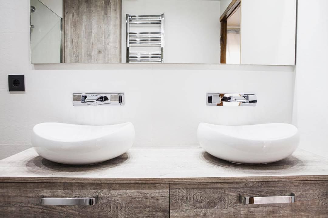 Dos lavabos ovalados blancos sobre un tocador de madera con grifería cromada. Espejo y toallero al fondo.