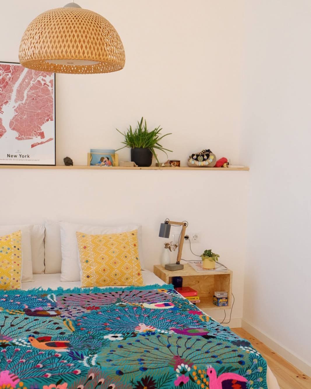 Dormitorio con cama, estantería con plantas y lámpara tejida. Manta verde azulado y almohadas amarillas.