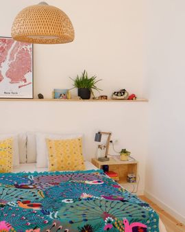 Dormitorio con cama, estantería con plantas y lámpara tejida. Manta verde azulado y almohadas amarillas.