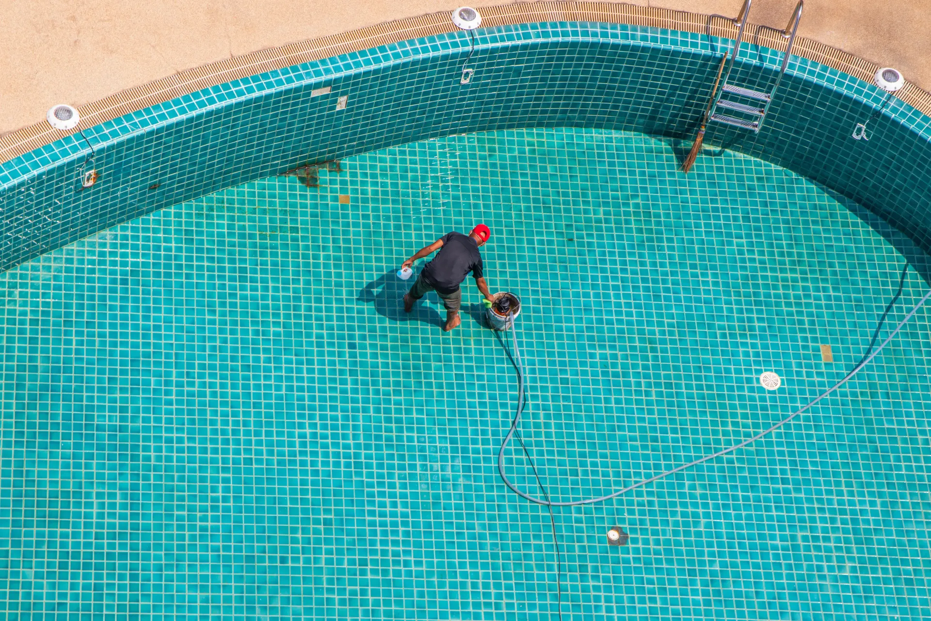 Persona reparando el suelo de baldosas de una piscina vacía. Vista aérea. Azulejos verde azulado y casco rojo.