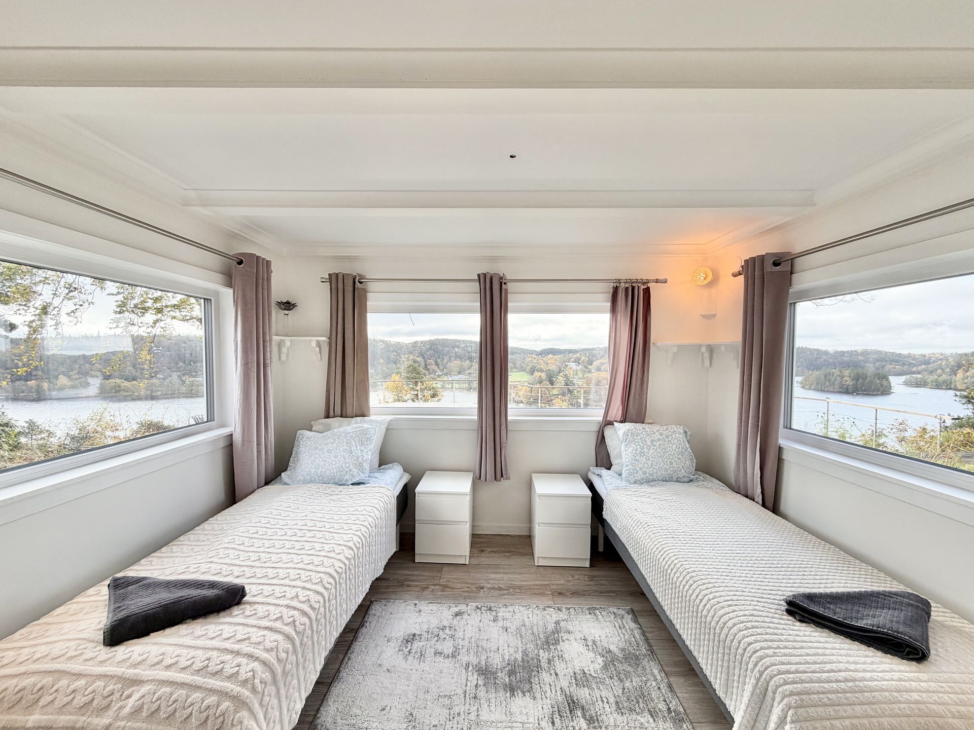 Twin beds in a room with large windows overlooking a lake, white walls, and gray rug.