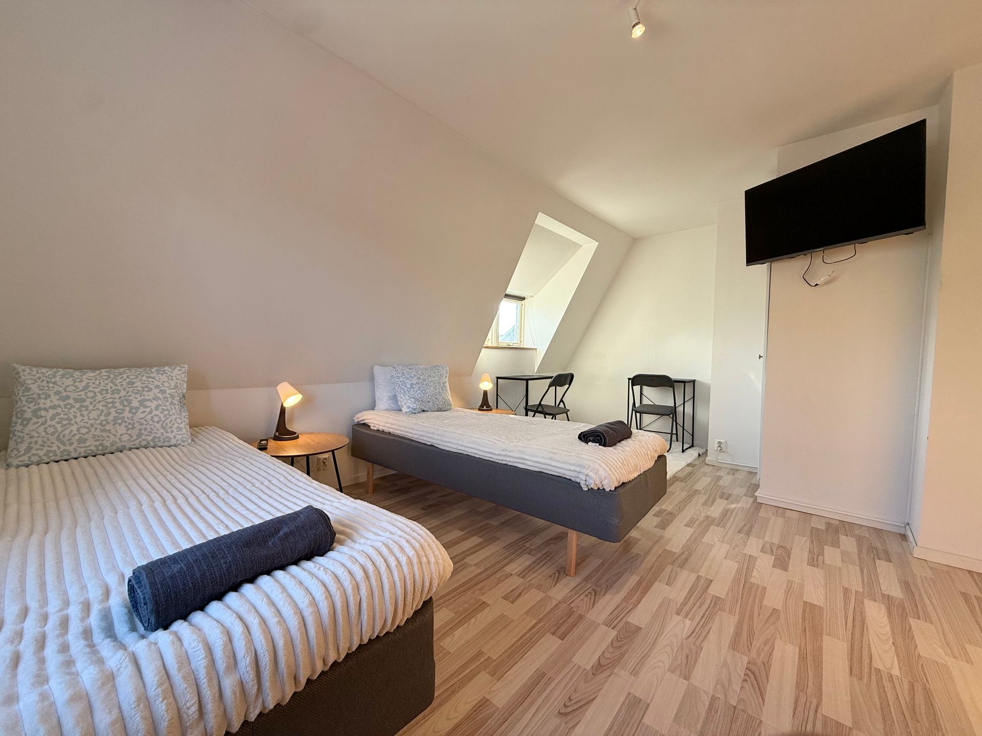 Bedroom with two twin beds, small tables, and a wall-mounted TV. Angled ceiling and wood-look flooring.
