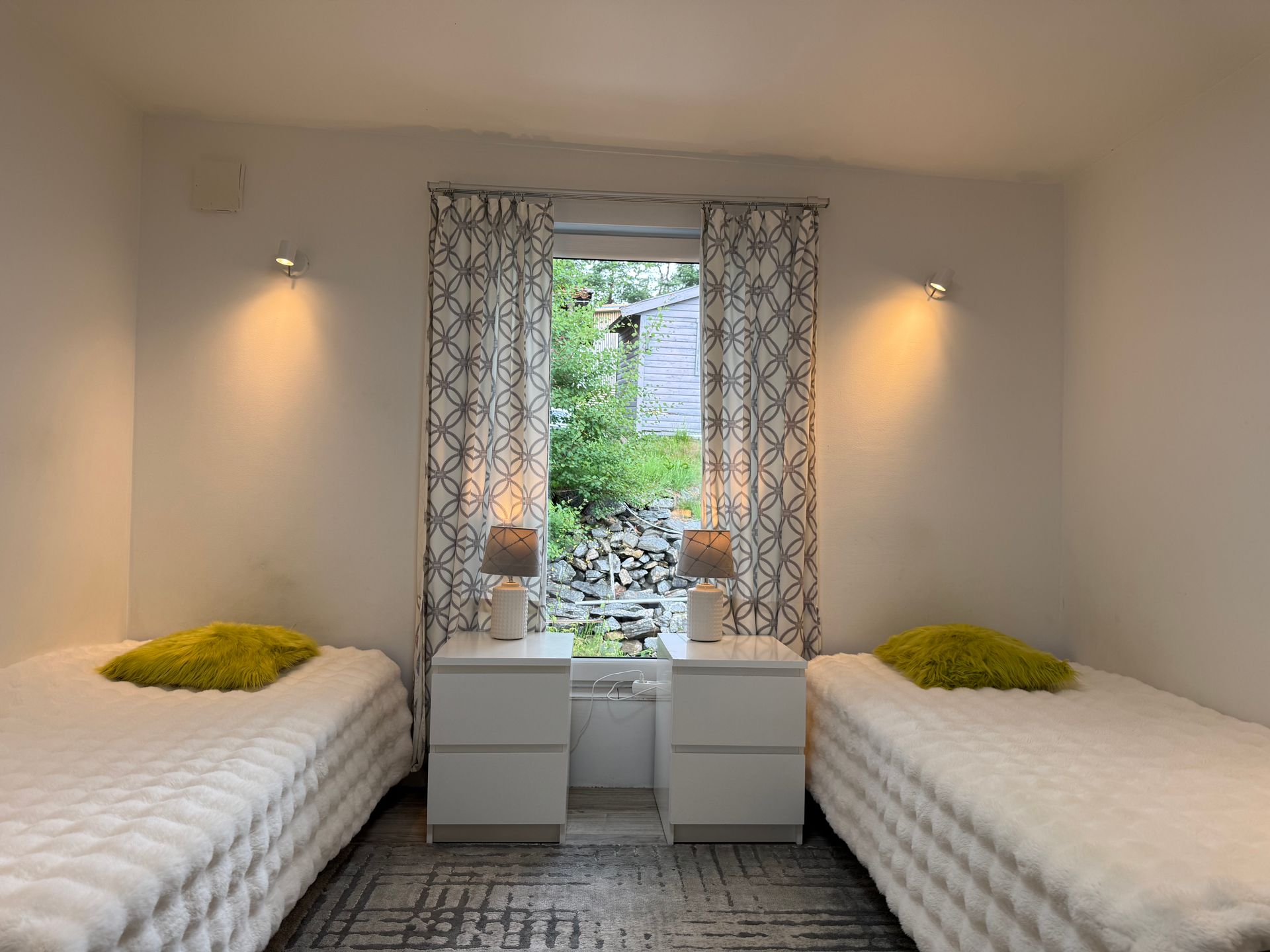 Two beds with white bedding, flanked by nightstands and lit by wall sconces, window with patterned curtains.