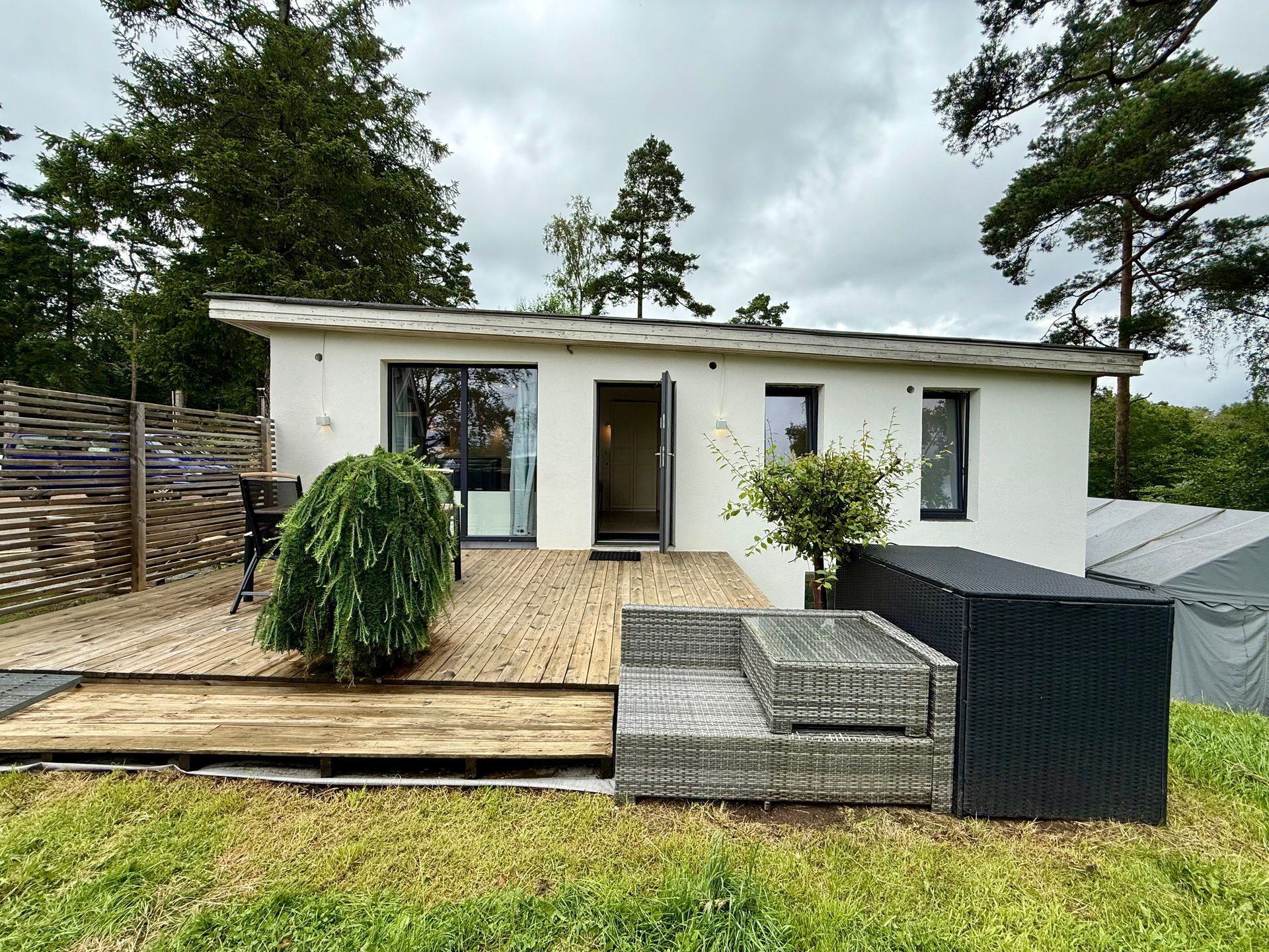 White cabin with a wooden deck, a small weeping tree, and black outdoor furniture on a grassy lawn.