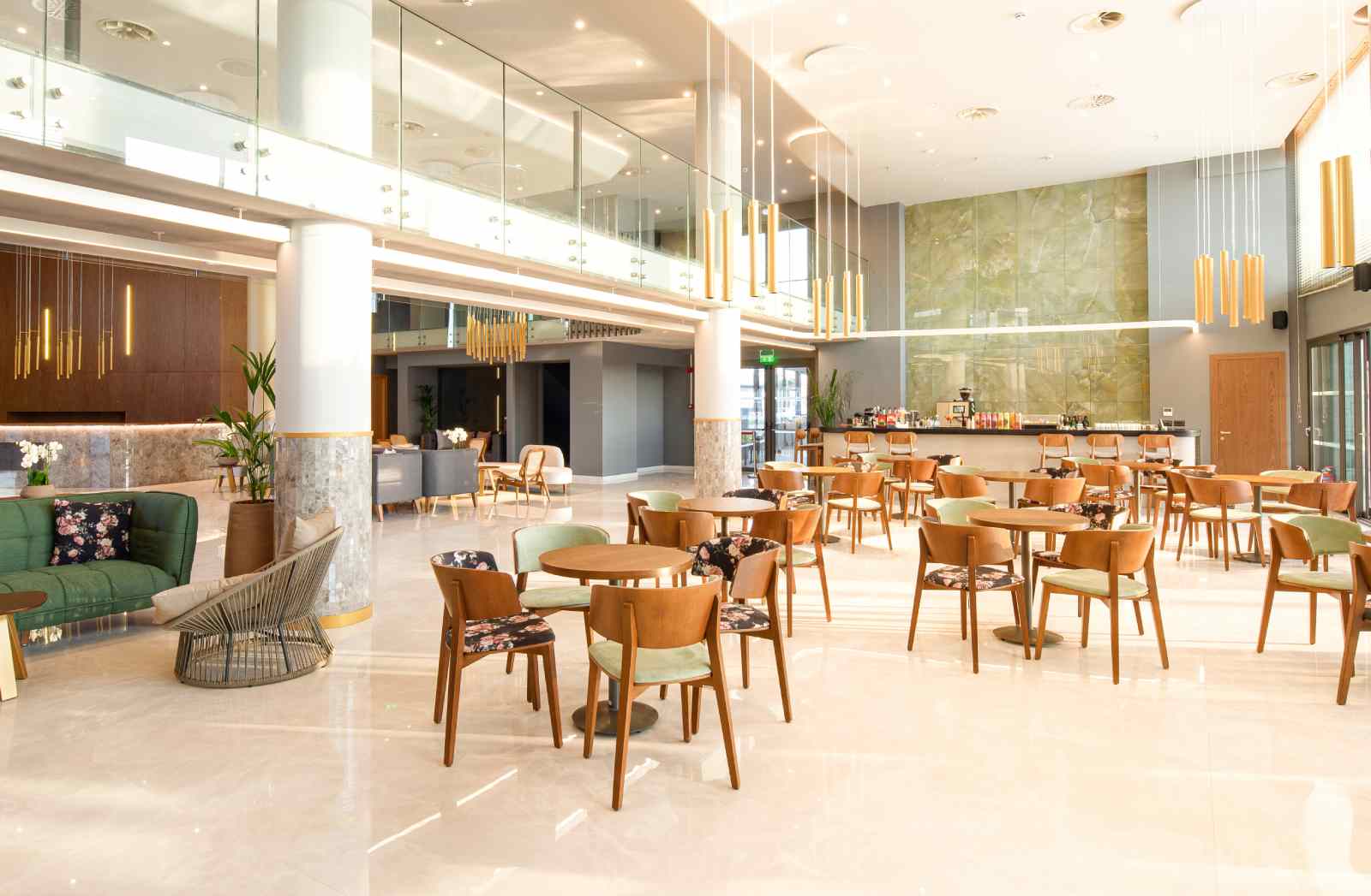 Bright hotel lobby with tables, chairs, bar, and seating area. Large windows, marble floors, and decorative lighting.