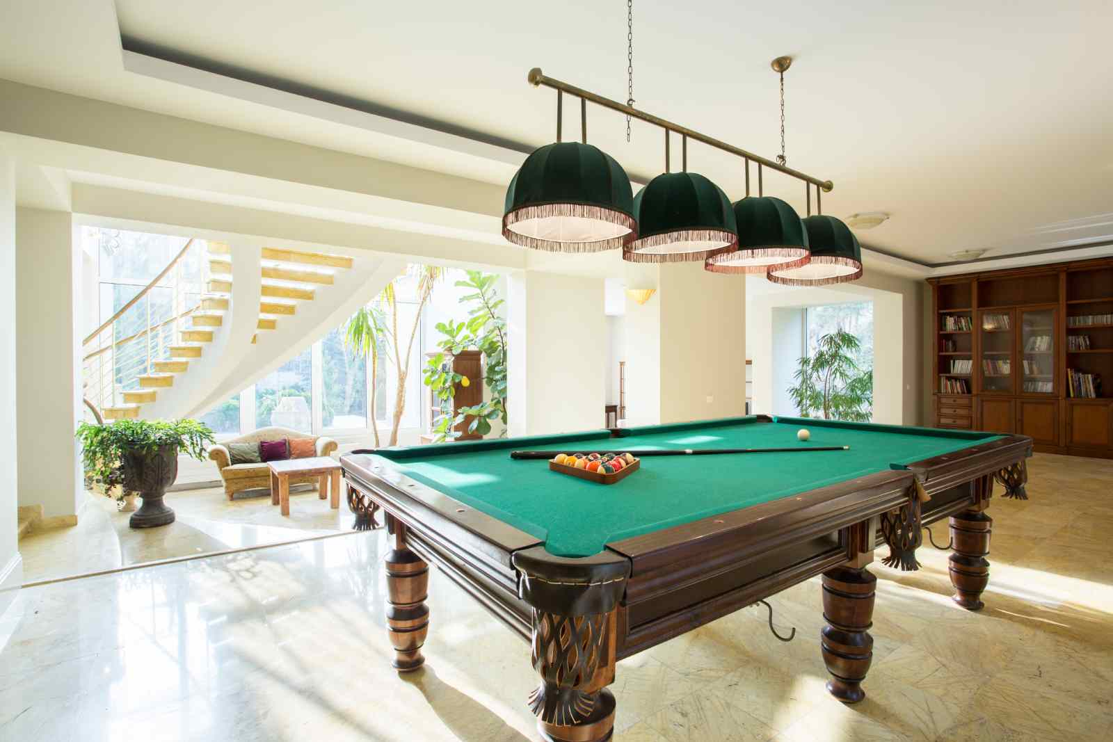 Pool table in a bright room with green felt, pendant lights, and a view of a staircase and trees.