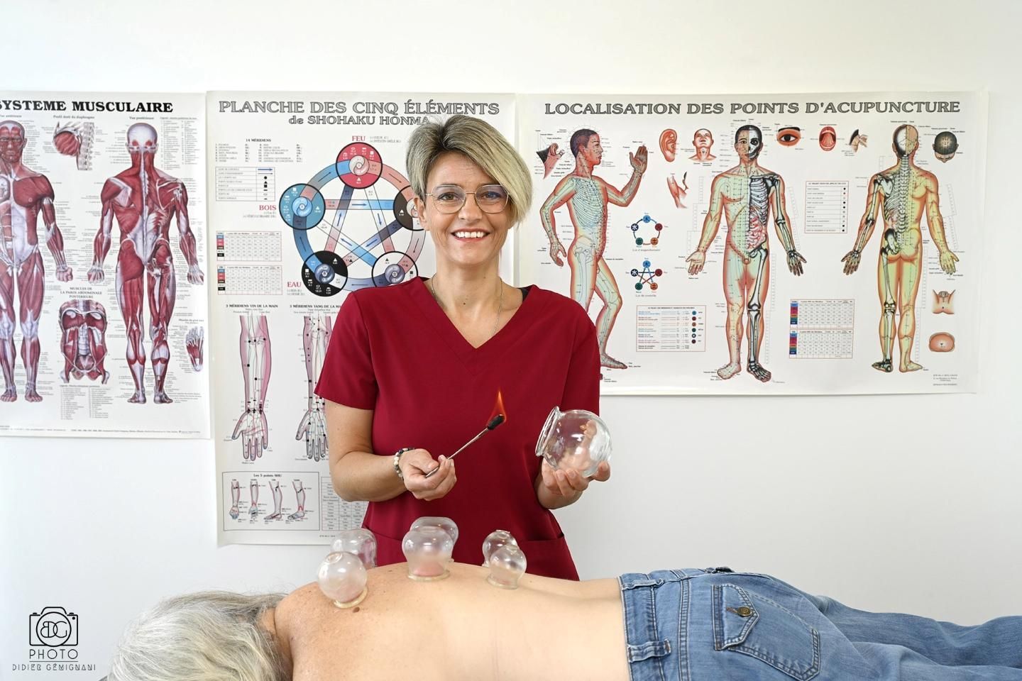 Une femme en haut rouge prodigue un soin à une personne allongée sur le ventre dans une clinique, devant des planches d'anatomie accrochées au mur.