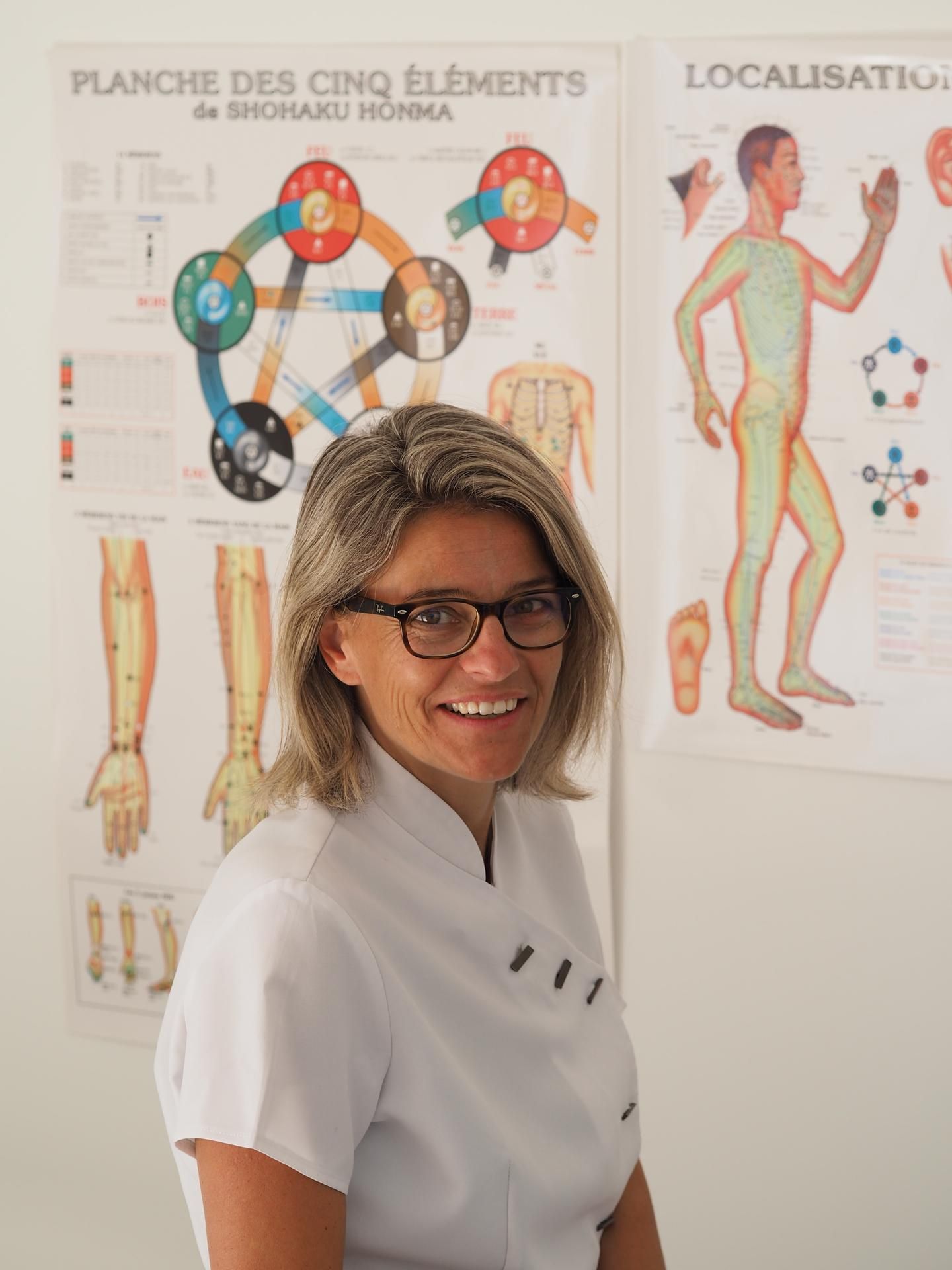 Femme souriante en blouse blanche et lunettes devant des planches anatomiques murales.