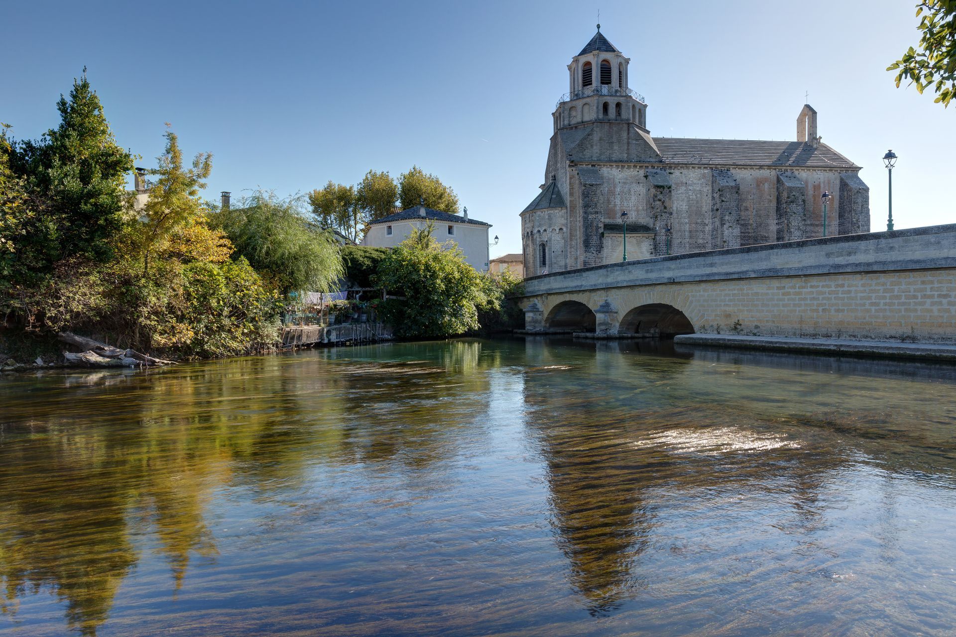 Église en pierre au bord d'une rivière sous un ciel bleu, avec des arbres et un pont se reflétant dans l'eau