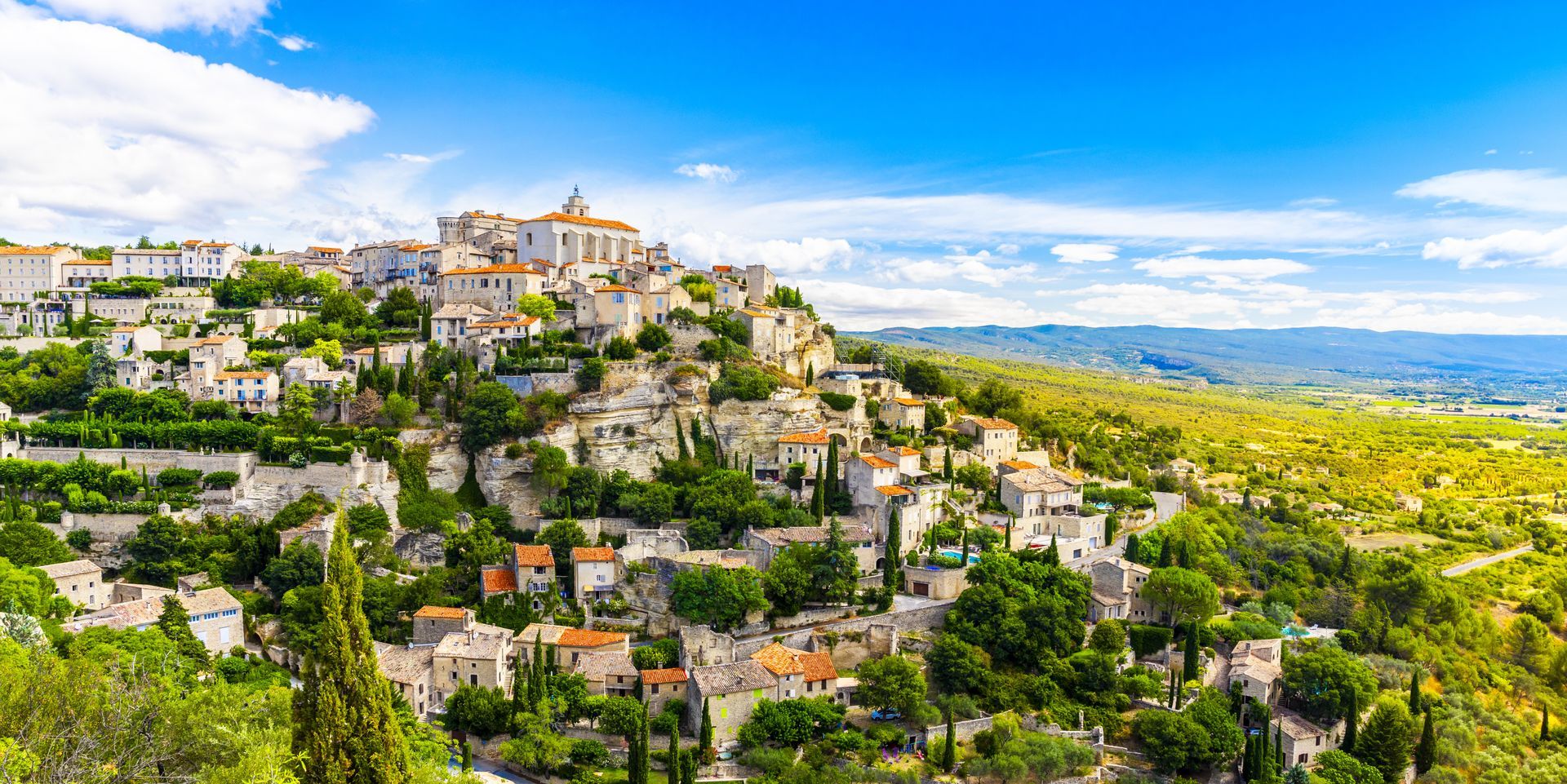 Village perché sur une colline, avec des bâtiments en pierre blanche au milieu de collines verdoyantes sous un ciel d'un bleu éclatant