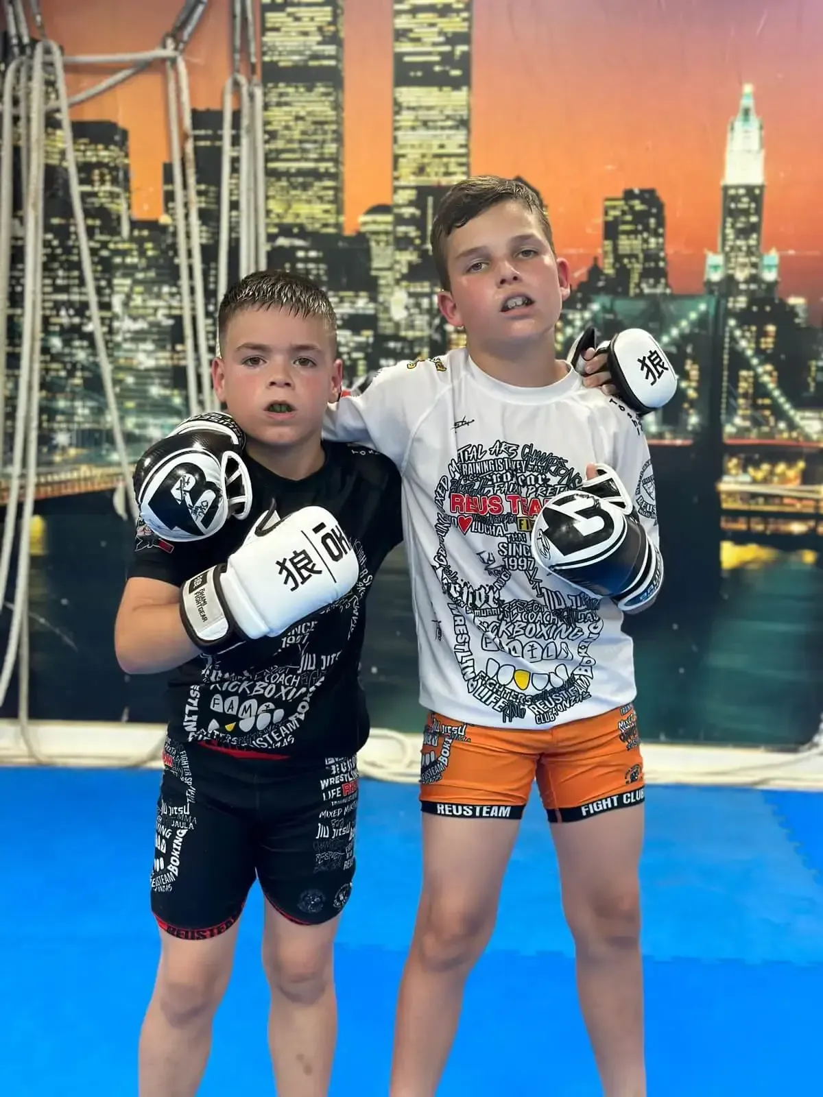 Dos niños con guantes de boxeo posando en un gimnasio con el perfil urbano de la ciudad de fondo.