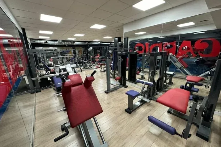 Interior de gimnasio con máquinas de ejercicio rojas y negras y paredes con espejos.