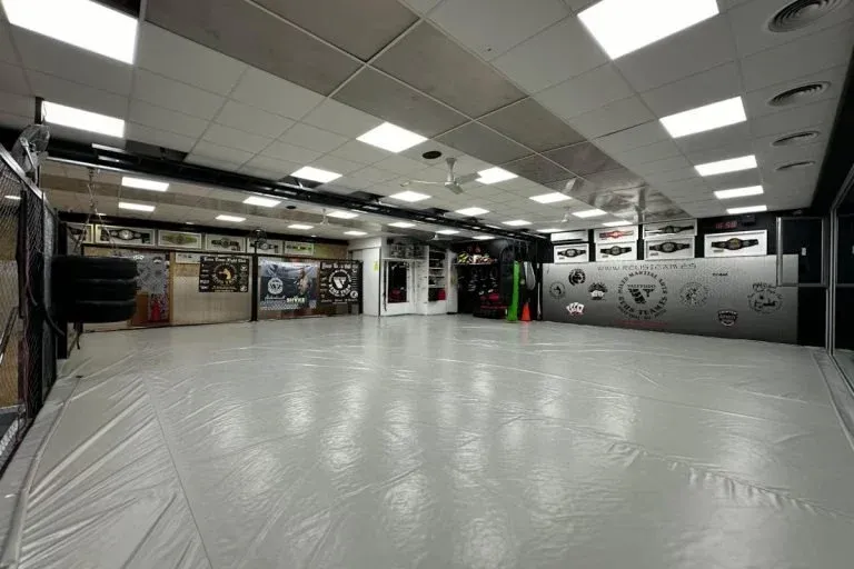 Amplia sala de entrenamiento de artes marciales con colchonetas blancas, luces fluorescentes y carteles en las paredes.