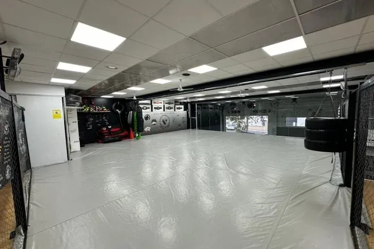 Garaje o taller interior vacío con suelo blanco, luces fluorescentes y un vehículo negro a la izquierda.