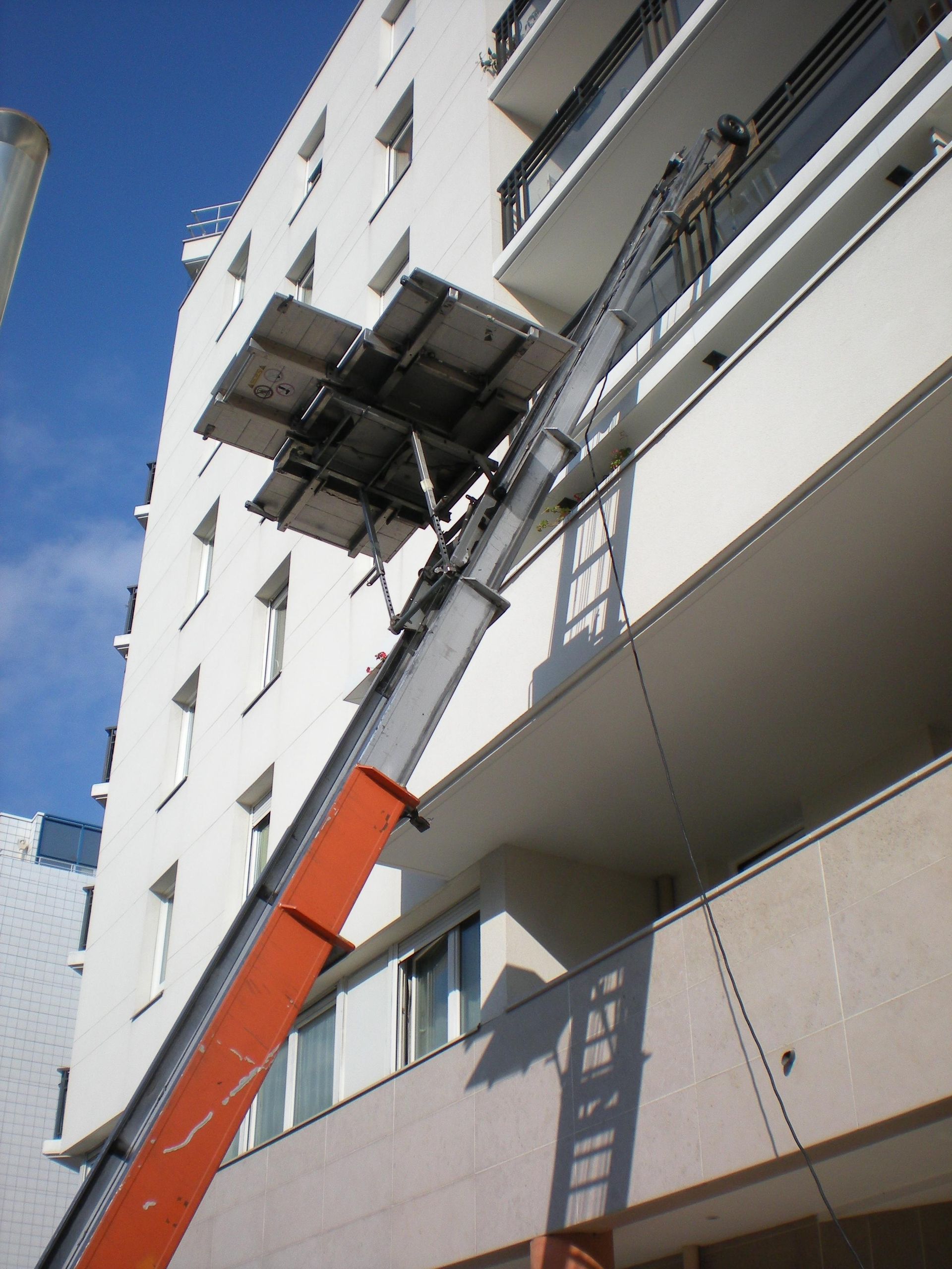 Un chariot élévateur orange soulève une plateforme jusqu'au balcon d'un immeuble blanc pour y déplacer des objets.