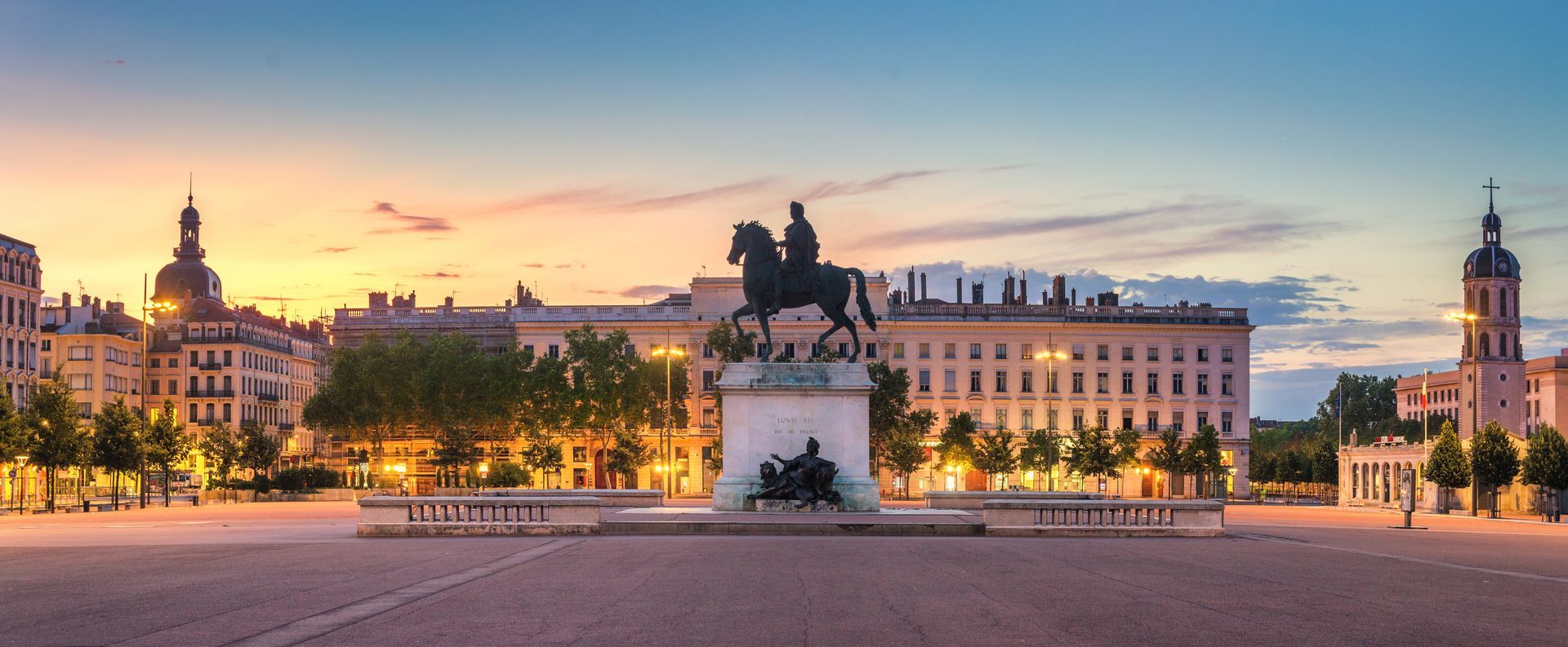 Statue équestre monumentale sur une place de Lyon, en France, au crépuscule. Des bâtiments se dressent à l'arrière-plan.