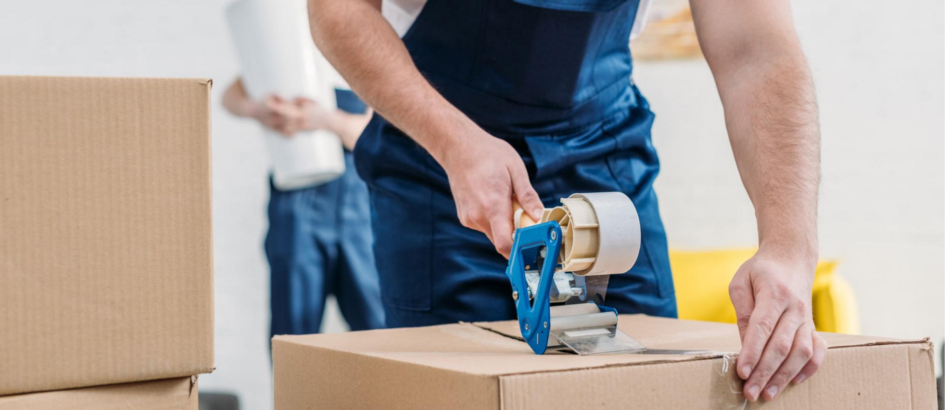 Une personne scotche une boîte en carton, une autre tient un rouleau de papier, à l'intérieur.