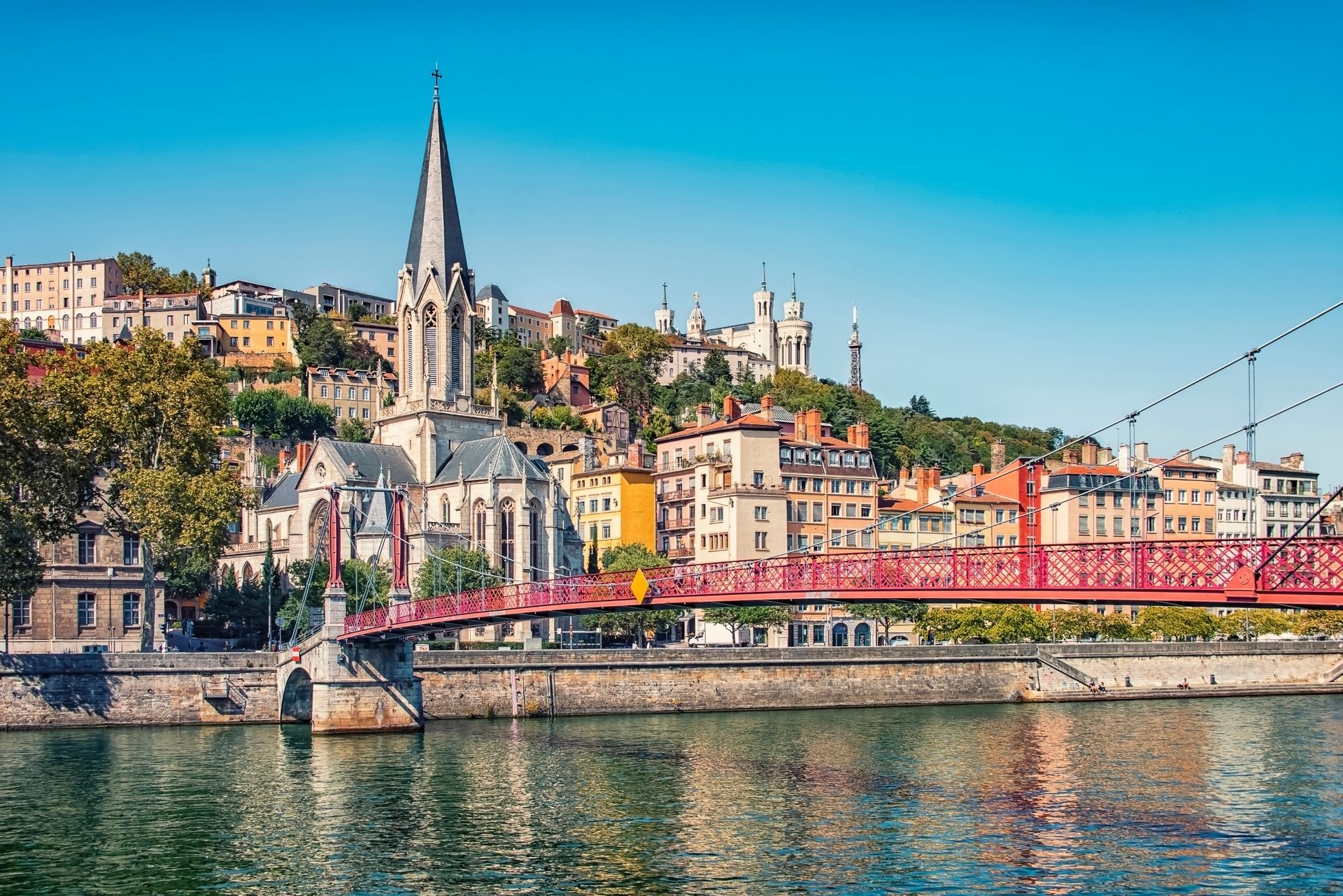 Lyon en France : paysage urbain avec un pont rouge, un clocher et des bâtiments à flanc de colline, sous un ciel bleu clair.