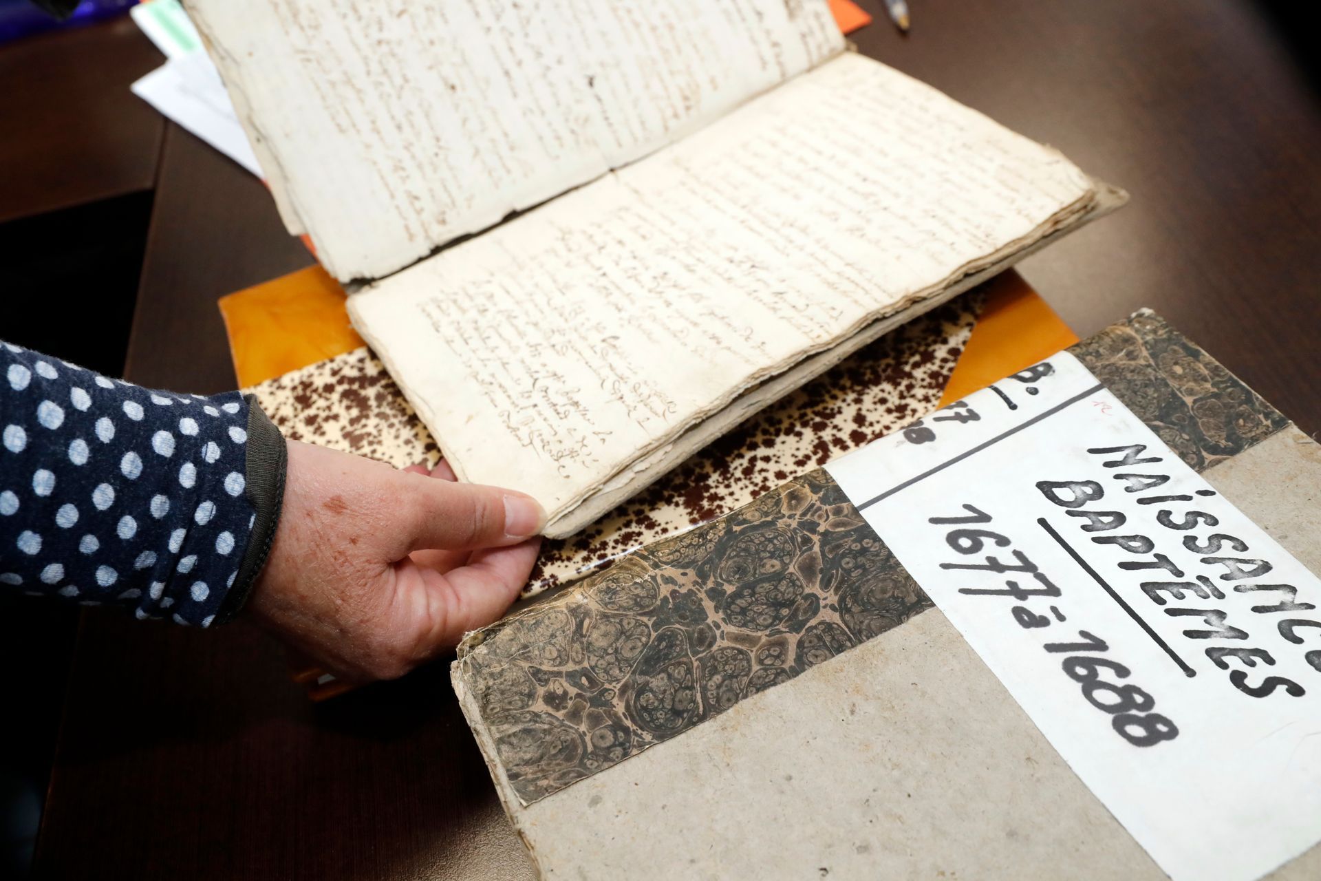 Une personne tient ouvert un vieux livre à l'écriture manuscrite, posé sur deux autres livres.