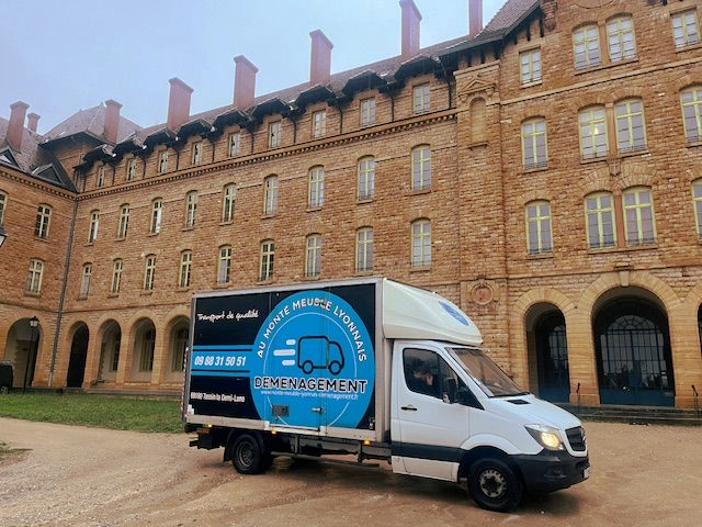 Camion de déménagement blanc stationné devant un grand bâtiment en briques. Logo et inscription bleus.