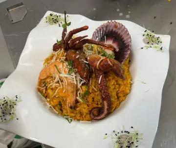 Paella de mariscos en un plato blanco, cubierta con camarones, pulpo y una vieira, adornada con brotes.