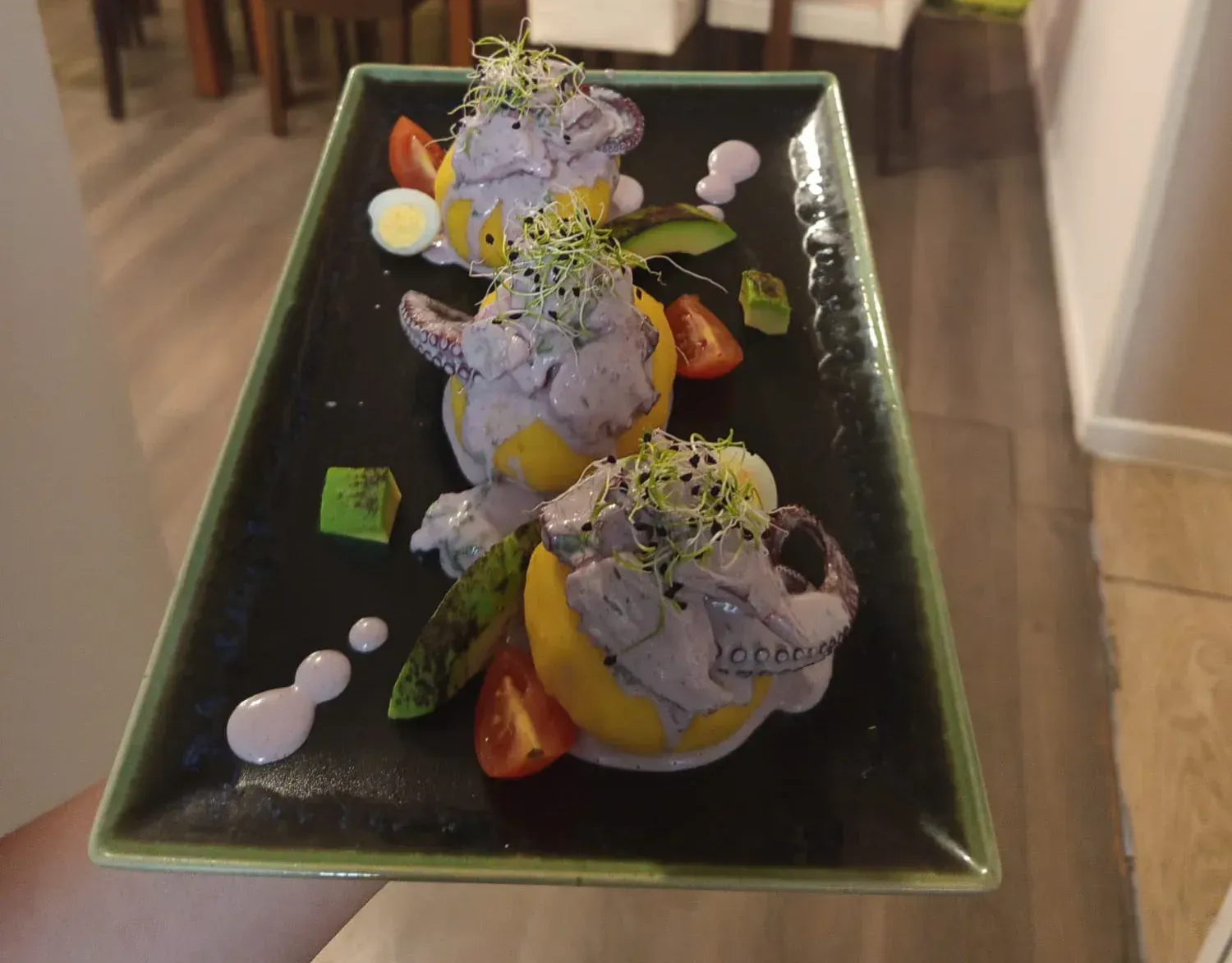 Tres porciones de comida esféricas cubiertas con salsa morada, brotes y guarniciones, servidas en un plato rectangular negro.