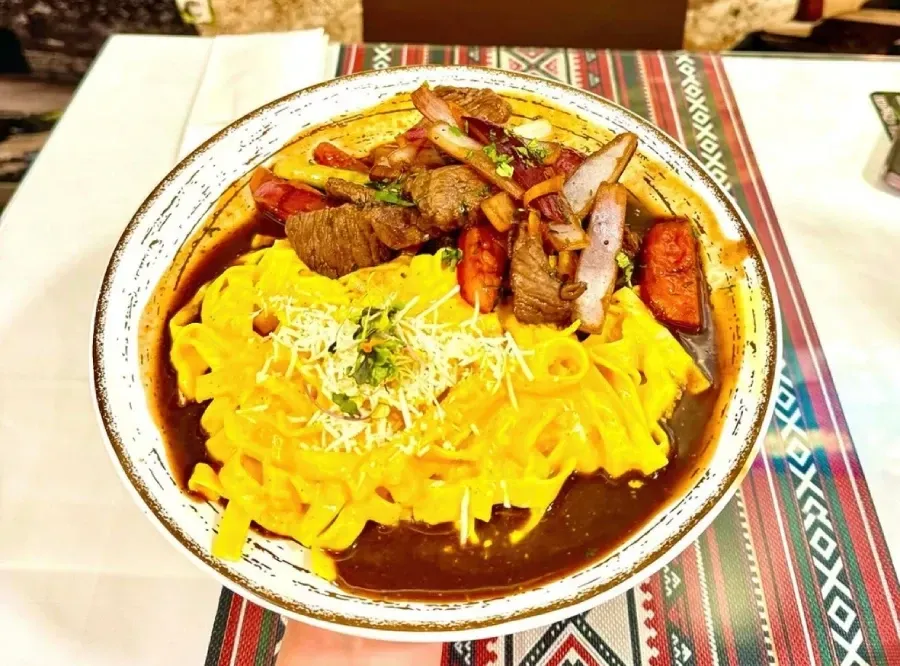 Plato de pasta con carne, salchicha y queso sobre un mantel estampado.