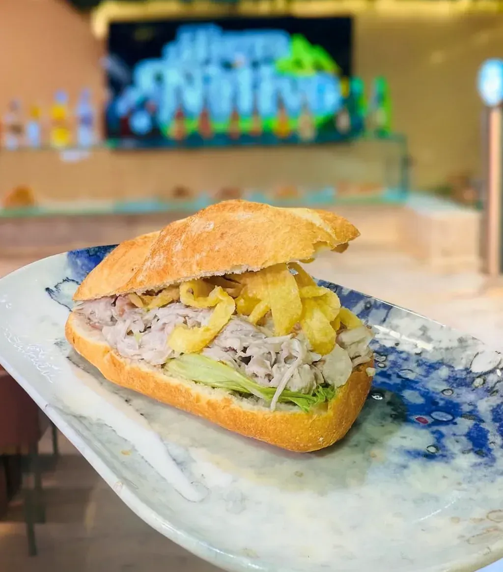 Sándwich en un plato decorativo, abierto con carne, patatas fritas y lechuga. El fondo parece un bar.