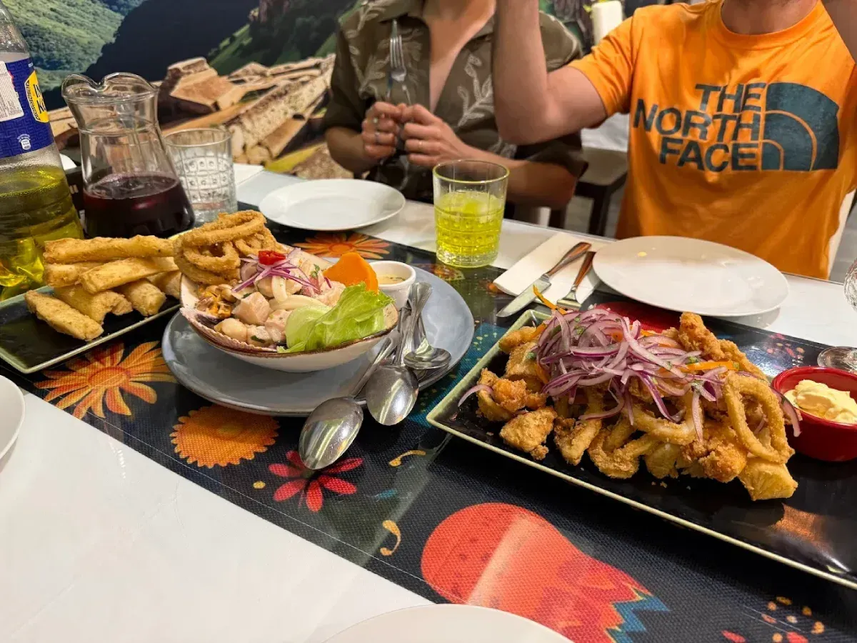 Mesa con platos de comida, incluyendo ceviche y calamares fritos, y gente sentada.