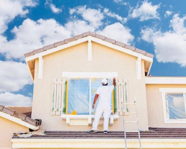 Un operaio dipinge l'esterno di una casa beige sotto un cielo azzurro.