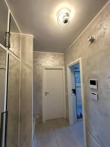 Stretto corridoio con pareti grigie, porta bianca, pavimento piastrellato e lampada a soffitto.