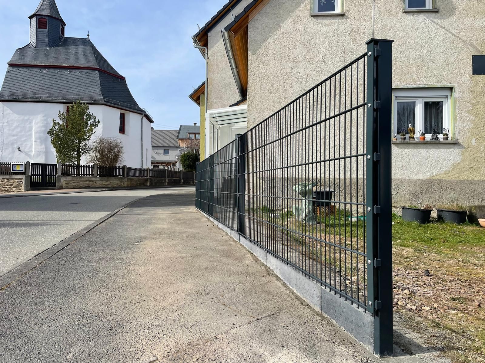 Schwarzer Metallzaun entlang eines Gehwegs, neben einem Haus und einer Kirche in einer Kleinstadt.