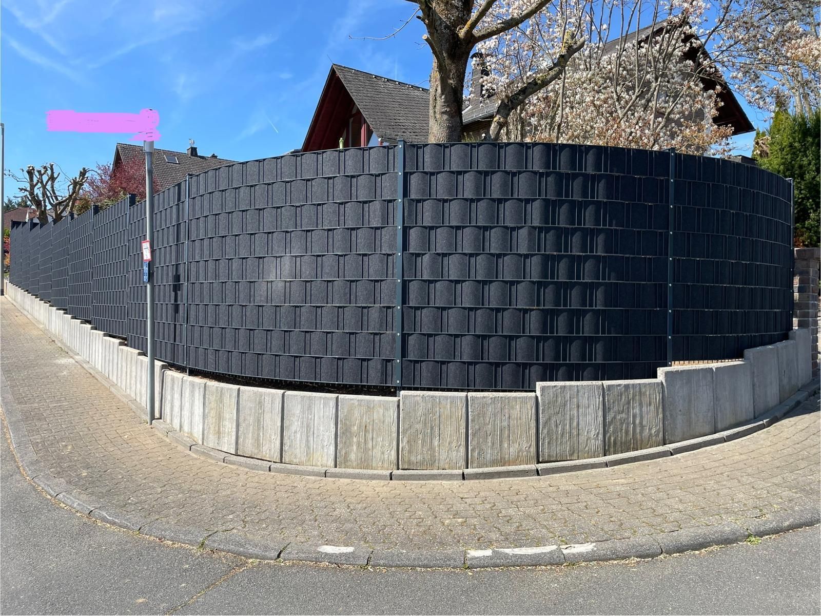 Ein dunkelgrauer, gewebter Sichtschutzzaun auf Betonblöcken begrenzt eine Straßenecke.