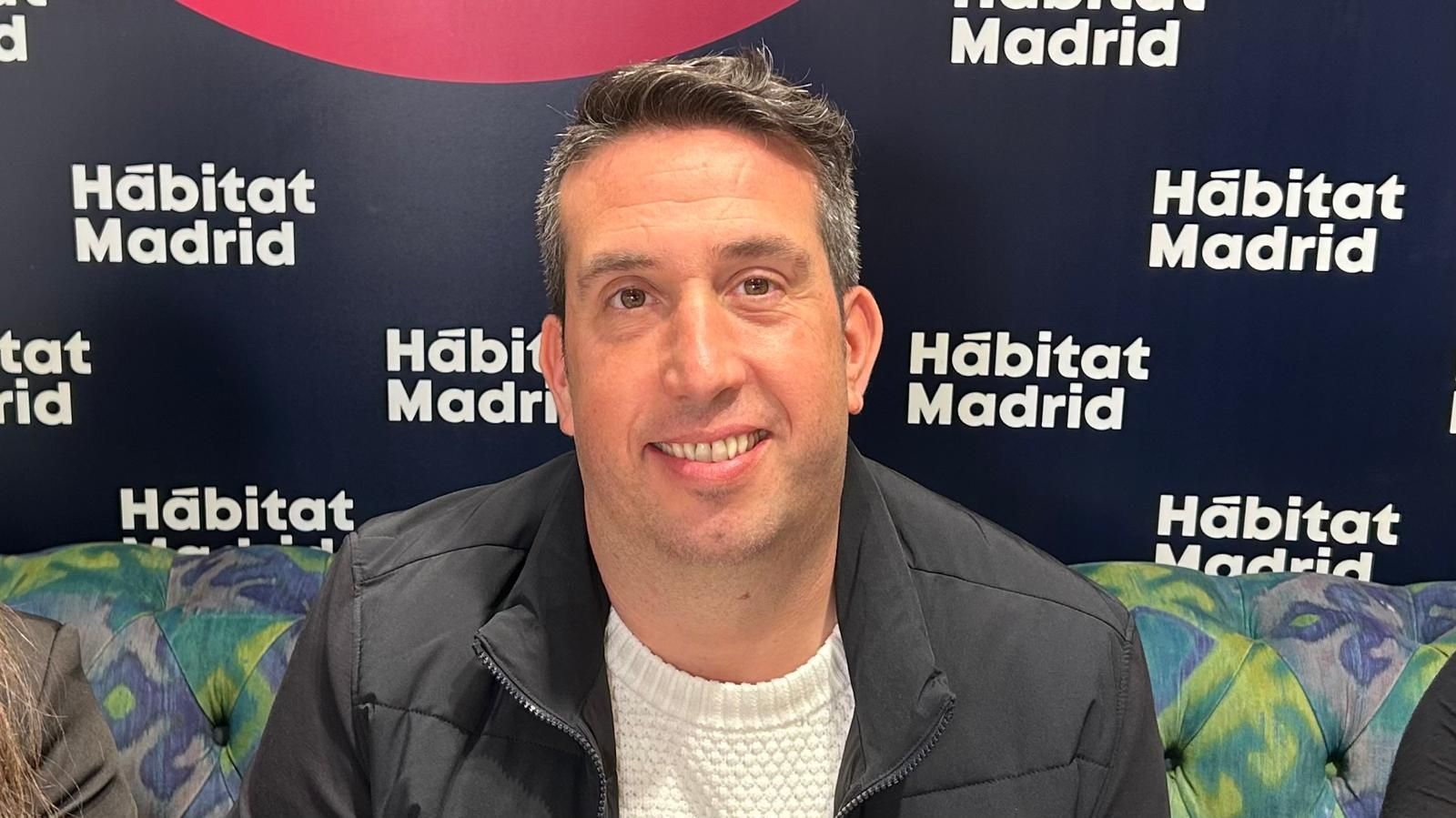 Hombre sonriente sentado frente a un telón de fondo de Habitat Madrid, con chaqueta oscura y jersey blanco.