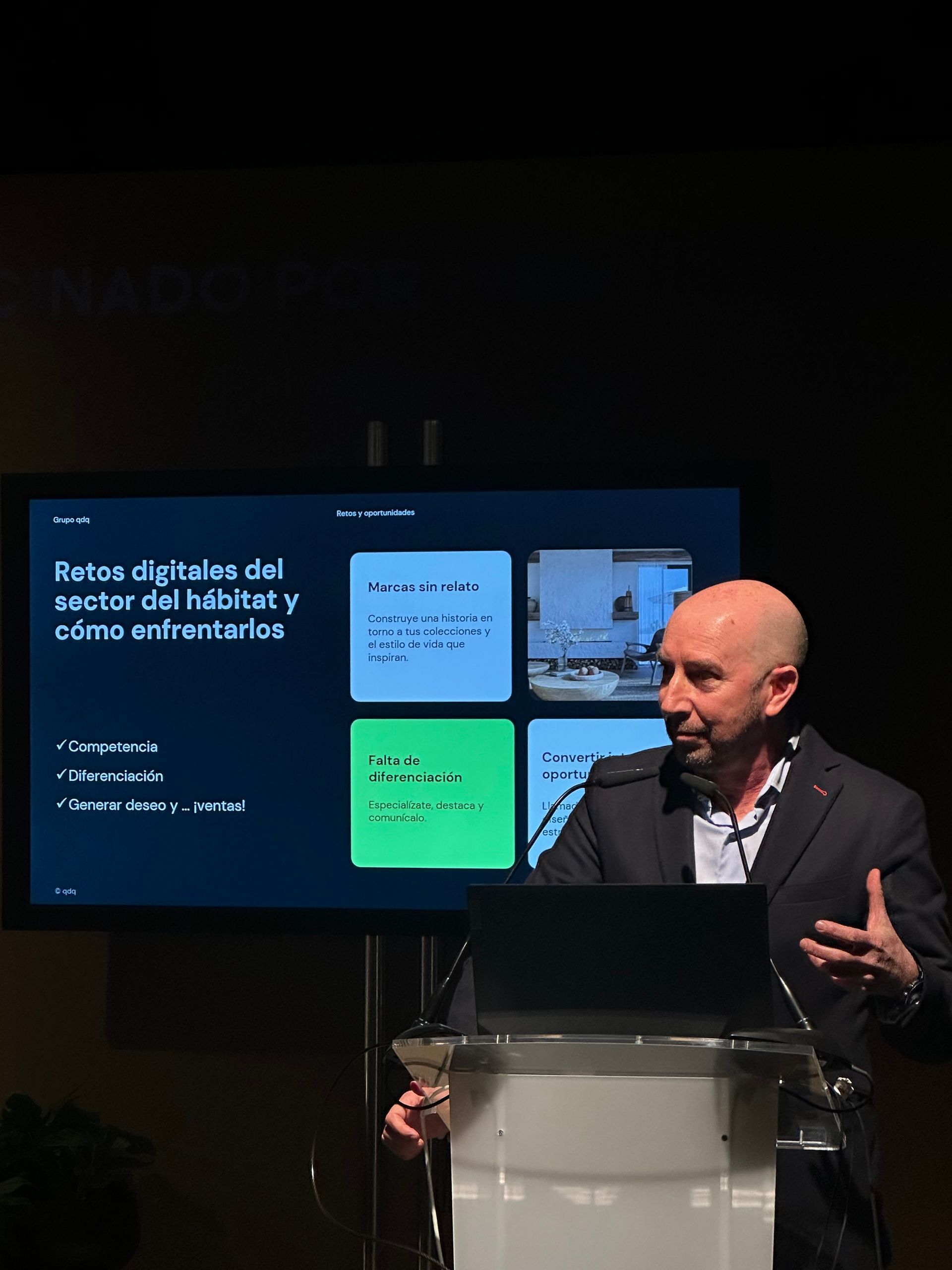 Orador en el podio presentando diapositivas en un escenario oscuro, con gráficos azules y verdes en la pantalla.