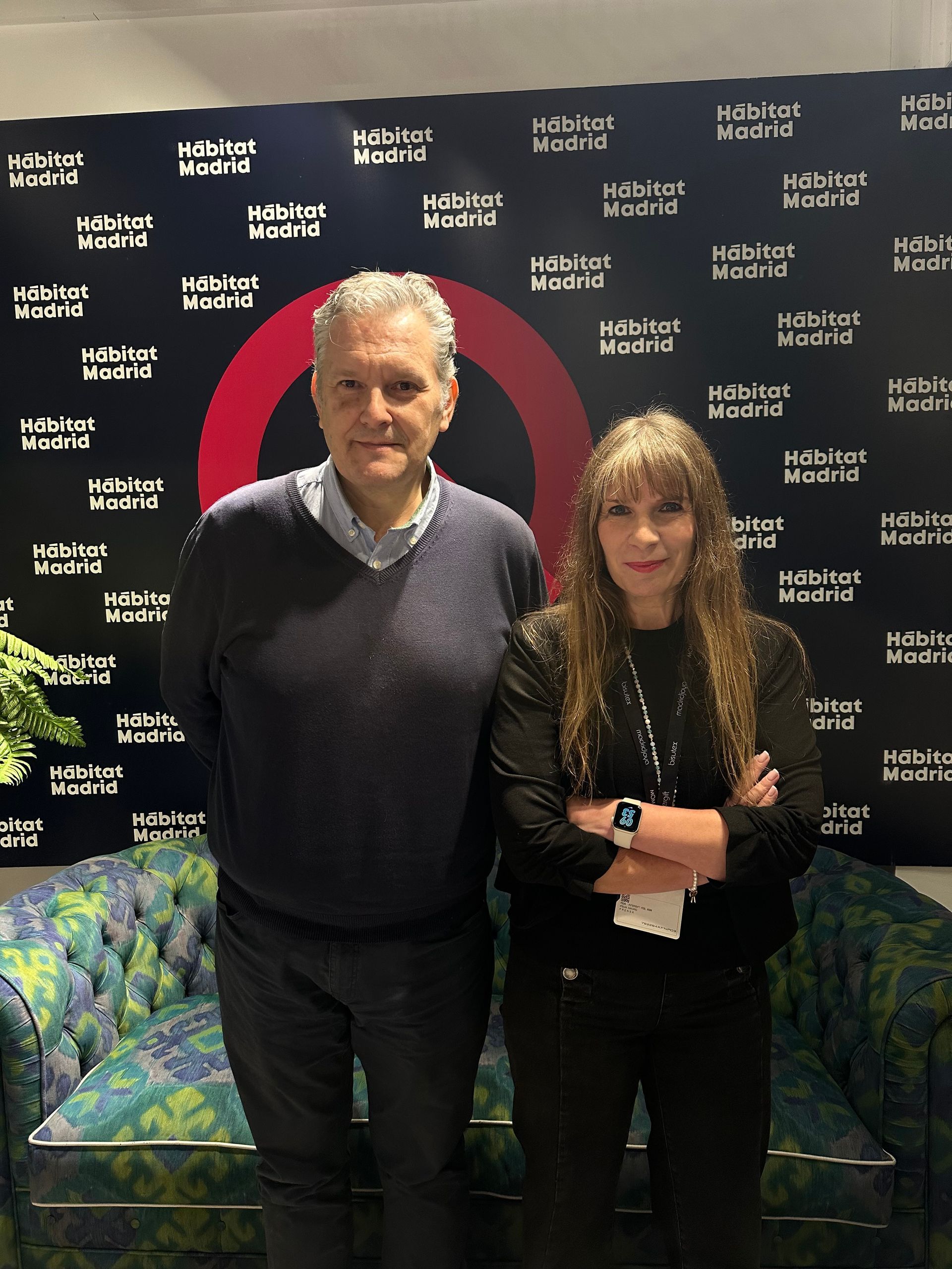 Dos personas de pie frente a un fondo con el logotipo de la marca, posando con los brazos cruzados