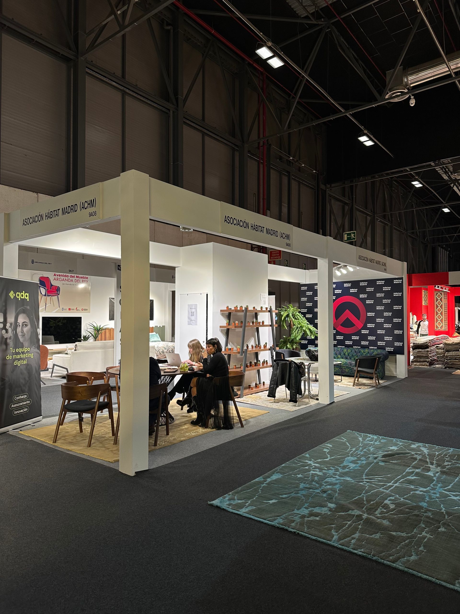 Stand de feria comercial con toldo blanco, mesas, sillas y personas sentadas dentro de una sala de exposiciones.