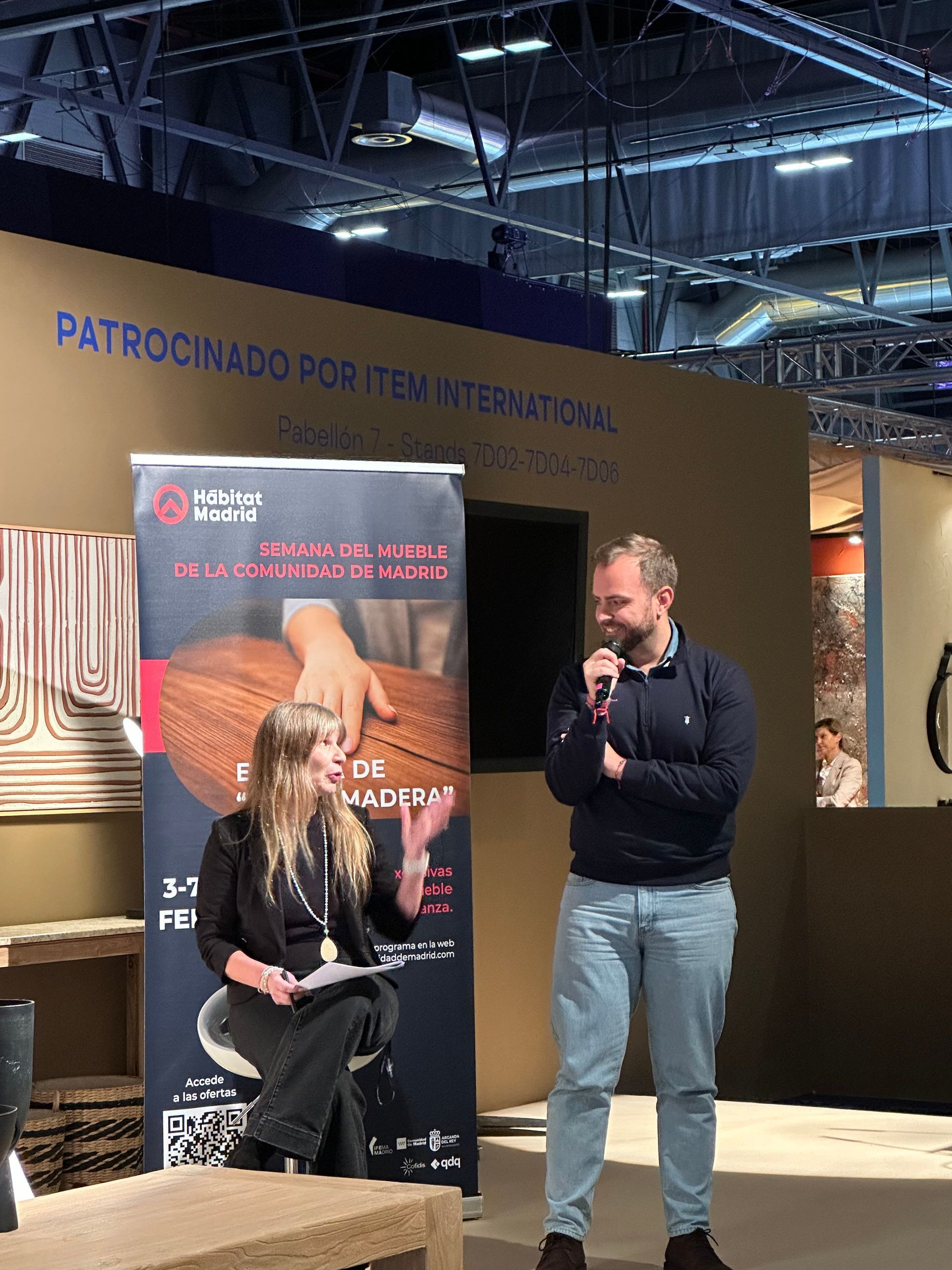 Dos personas hablando en un escenario durante un evento en un espacio cerrado, con una pancarta publicitaria detrás de ellas.