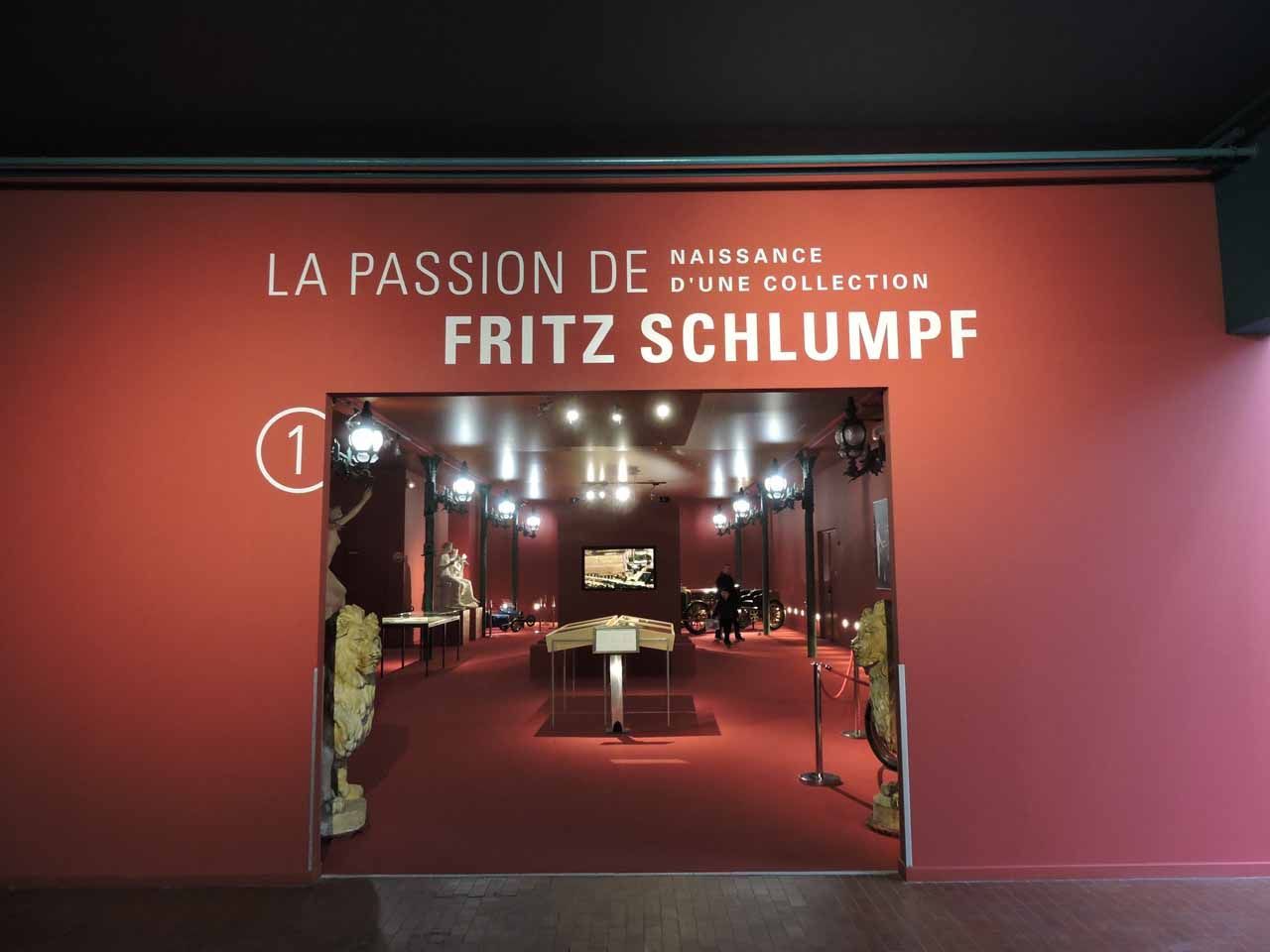 Entrée d'une exposition de musée, avec des murs rouges et un panneau indiquant « La Passion de Fritz Schlumpf ».