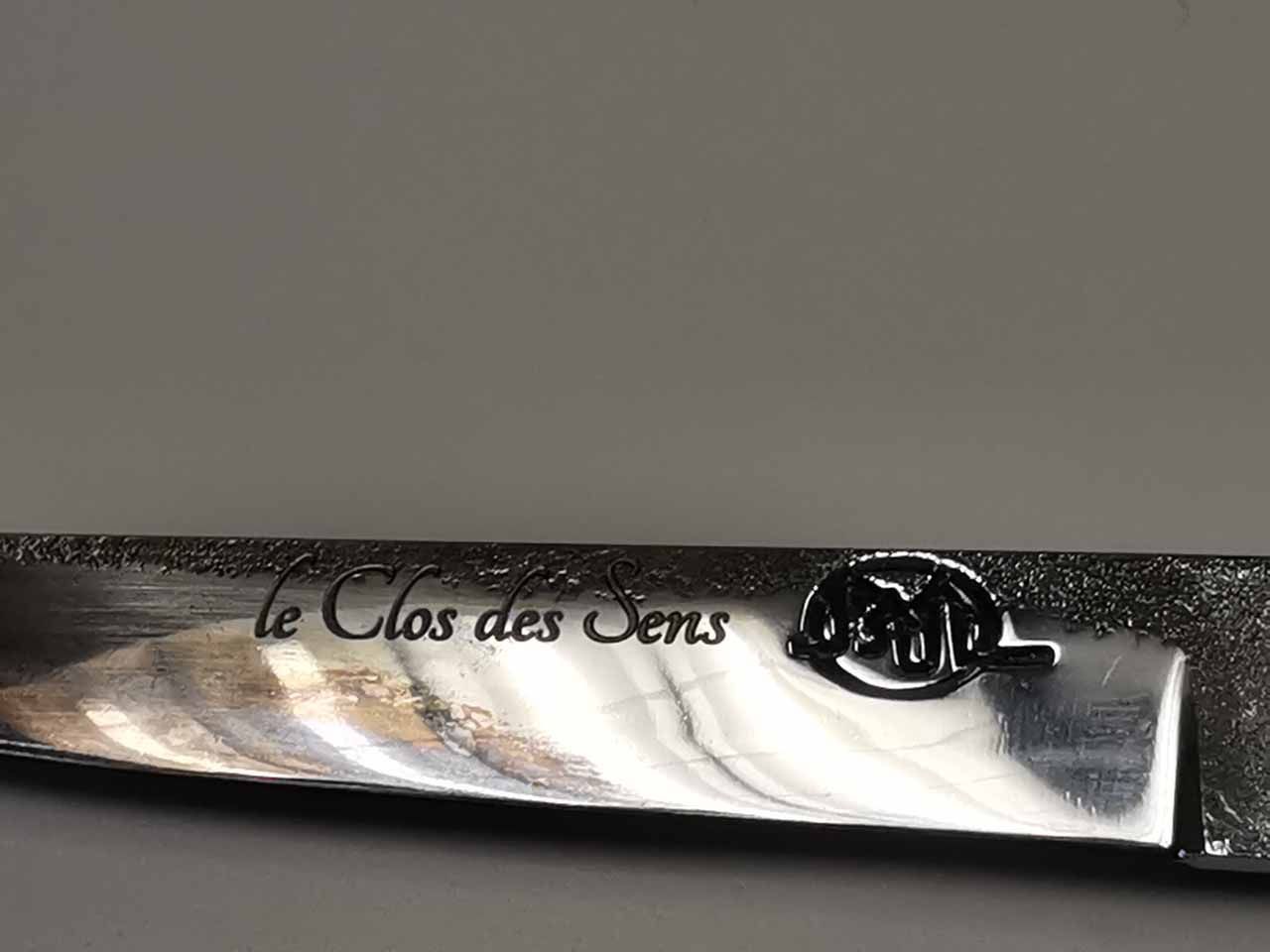Gros plan sur une lame de couteau gravée des mots « le Clos des Sens » et d'un emblème circulaire.