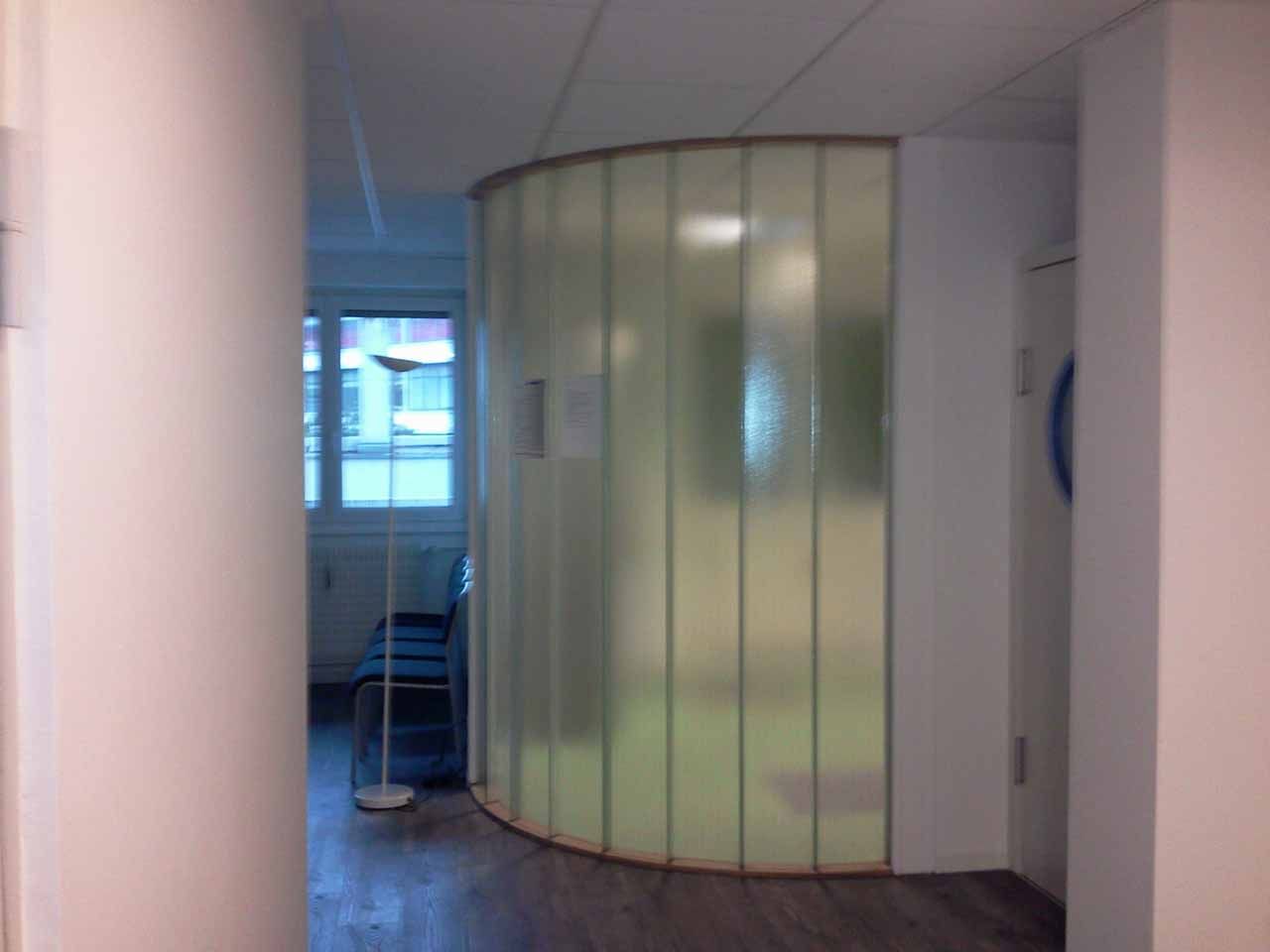 Couloir avec cloison incurvée en verre dépoli, menant à une salle d'attente avec chaises et fenêtre.