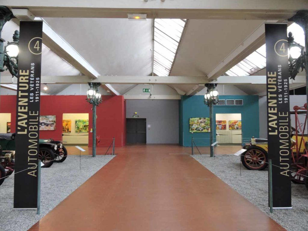 Couloir du musée avec des voitures classiques, des murs rouges et turquoise, et des panneaux indiquant « L'Aventure Automobile ».