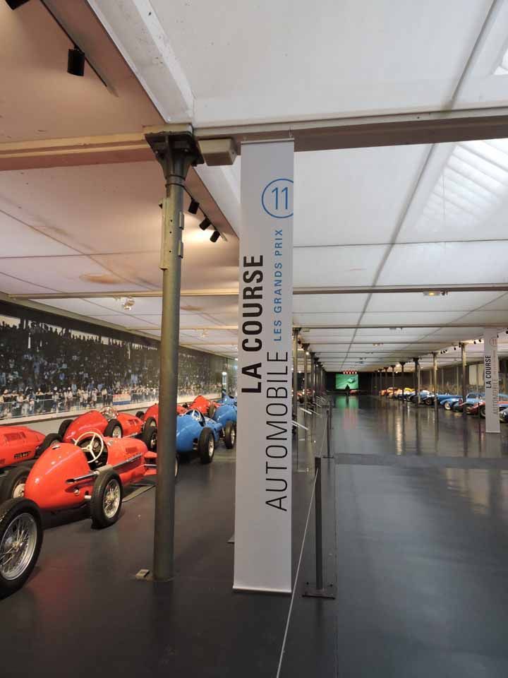 Exposition de voitures de course anciennes dans un long hall. Panneau : « La Course Automobile ».