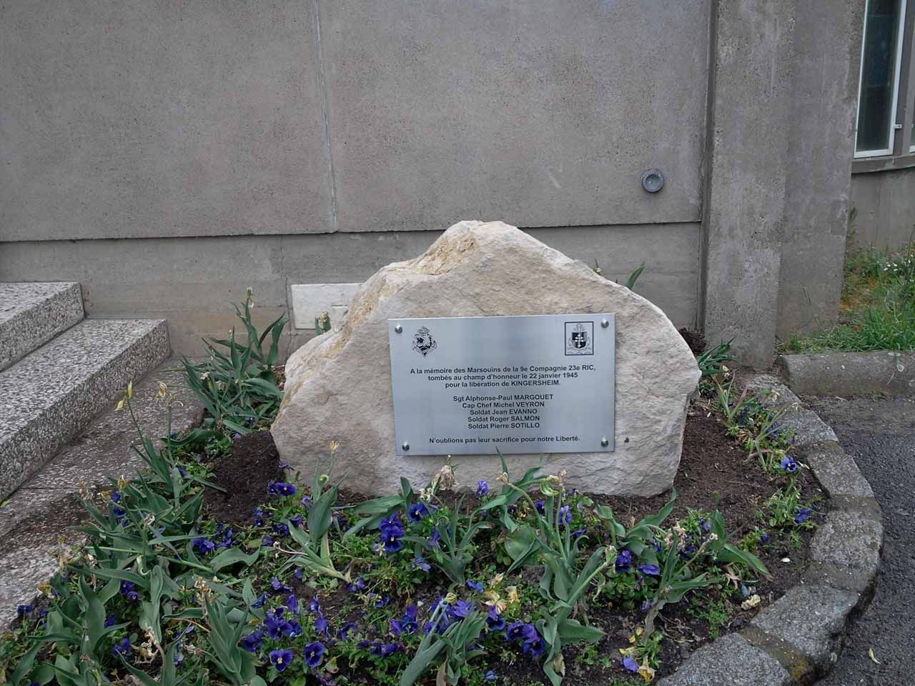 Plaque commémorative sur un gros rocher, entourée de fleurs, devant un bâtiment.