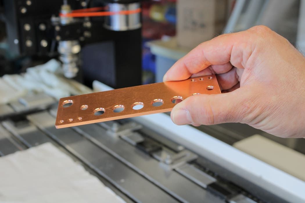 Main tenant une plaque de circuit imprimé en cuivre avec des trous de différentes tailles, probablement dans un atelier.