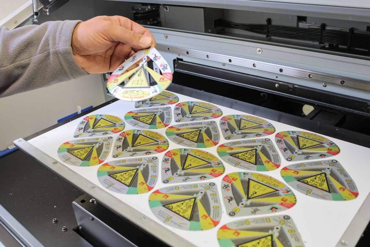 Une personne tient une étiquette imprimée à l'aide d'une imprimante grand format. Les étiquettes sont multicolores et comportent un symbole de danger.