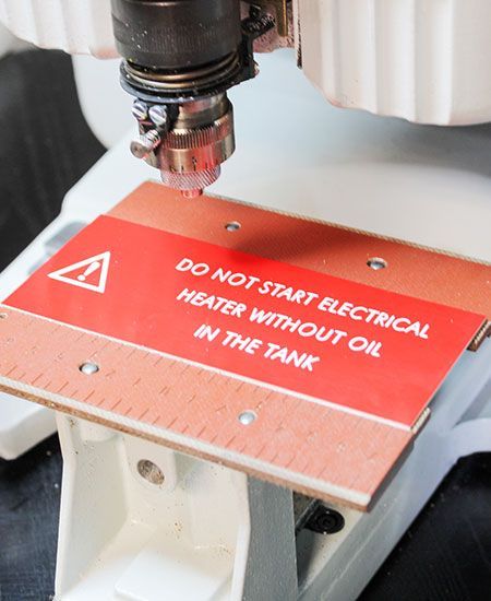 Machine CNC découpant un panneau d'avertissement rouge sur lequel on peut lire : « NE PAS DÉMARRER LE CHAUFFAGE ÉLECTRIQUE SANS HUILE DANS LE RÉSERVOIR. »